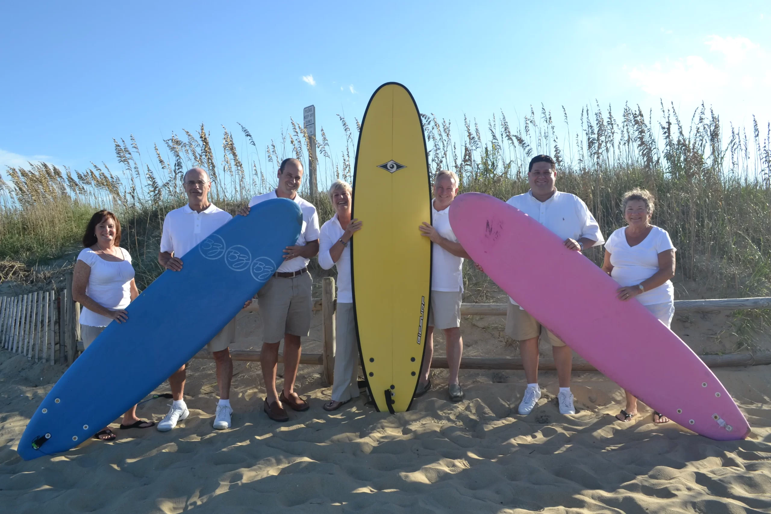 DSC_4080 Group of seven adults posing with colorful surfboards on a sandy beach - Siebert Realty Sales